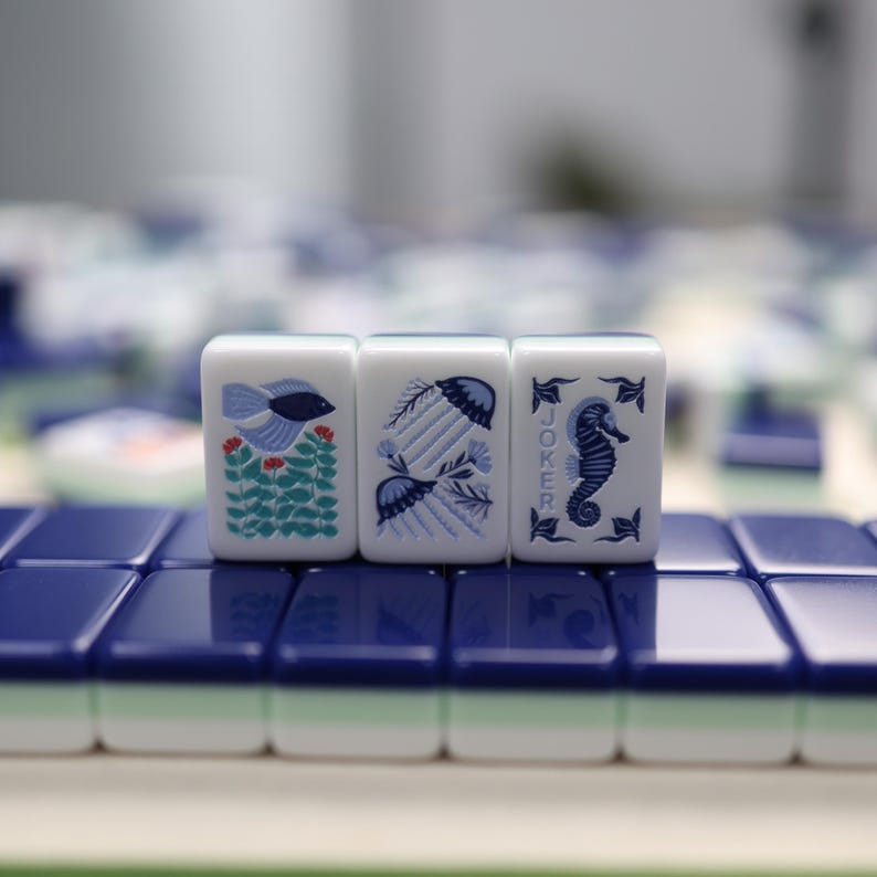 May include: Three white Mahjong tiles with blue illustrations of a fish, jellyfish, and seahorse. The tiles are arranged on a blue and white surface. The third tile has the word "JOKER" printed on it.