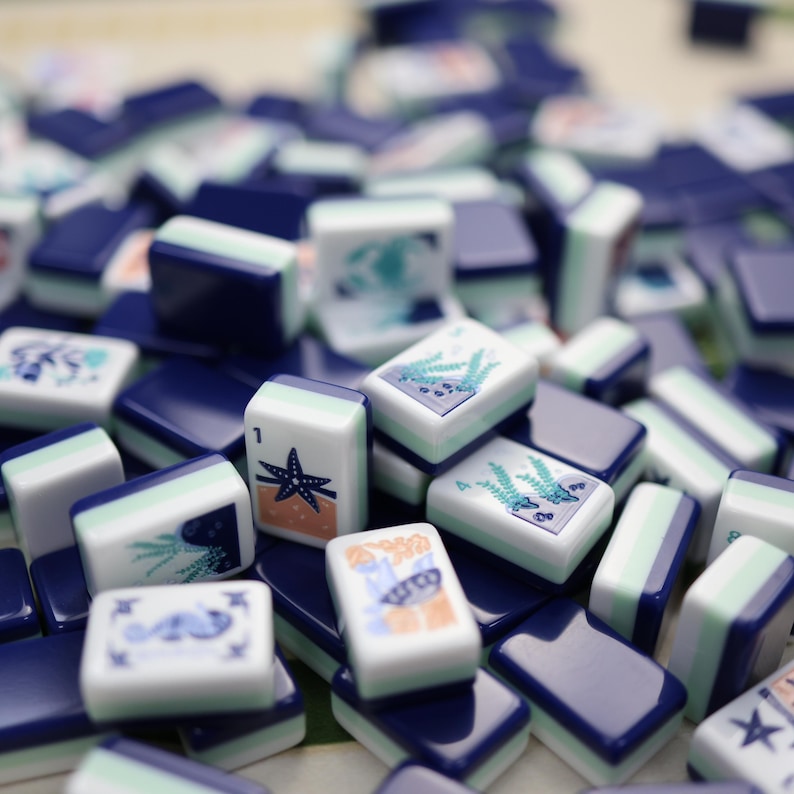 May include: A collection of blue and white Mahjong tiles with various sea-themed illustrations. Some tiles feature a starfish, while others display aquatic plants. The tiles are stacked together, creating a visually appealing arrangement.