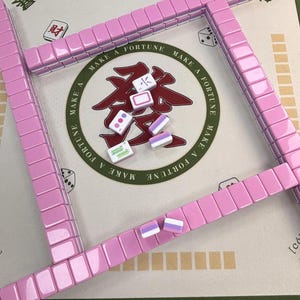 May include: A pink Mahjong set with tiles and a game board. The tiles are arranged in a square, with some tiles scattered in the center. The board has Chinese characters and the words "Make a Fortune".