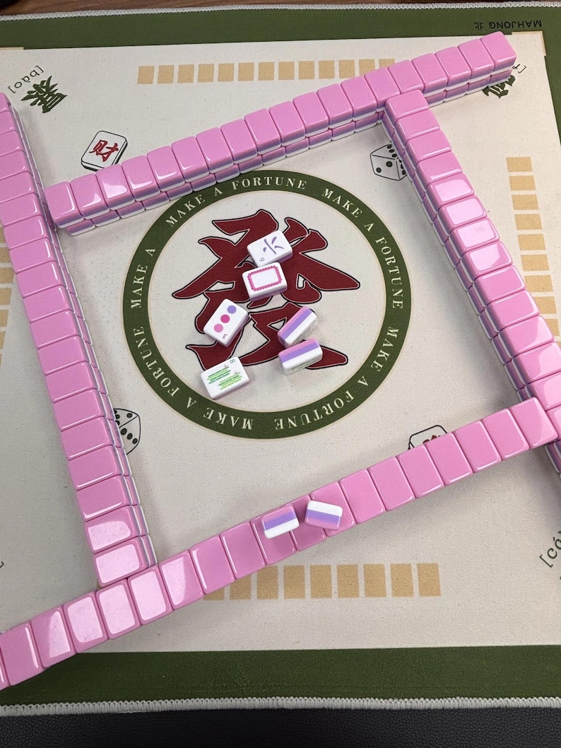 May include: A pink Mahjong set with tiles and a game board. The tiles are arranged in a square, with some tiles scattered in the center. The board has Chinese characters and the words "Make a Fortune".