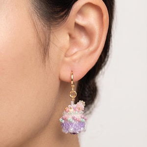 Könnte beinhalten: Ein goldener Creolen-Ohrring mit einem Cupcake-Anhänger aus Perlen. Der Cupcake besteht aus lila, rosa und weißen Perlen mit einer rosa Perlenblume oben. Der Ohrring wird an einem Ohr getragen.