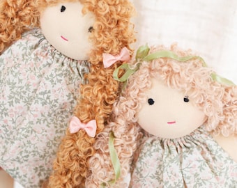 May include: Two handmade rag dolls with curly hair and fabric dresses. One doll has light brown hair with pink bows, the other has pink hair with green ribbon. Both dolls have floral print dresses and simple facial features.