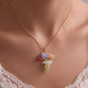 Peut inclure: Un collier doré avec un pendentif représentant un bouquet de fleurs en perles. Les fleurs sont roses, bleues et jaunes, nouées avec un nœud blanc. Le collier est porté autour du cou.