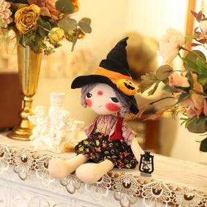 May include: A handmade Halloween witch doll with a black hat, orange band, and pumpkin accent. The doll has gray hair, rosy cheeks, and is wearing a floral dress and red vest. A small black lantern sits beside the doll.