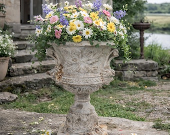 May include: A large, weathered stone urn overflowing with a vibrant mix of colorful flowers. The arrangement includes daisies, delphiniums, and other blooms in shades of white, yellow, pink, and purple. The urn is placed on a stone patio near a building.