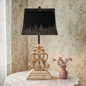 May include: A decorative table lamp with a black lampshade and a distressed gold-coloured base. The base features an ornate, scrolled design. The lamp sits on a round table with a lace tablecloth. A small pink vase with flowers is next to the lamp.
