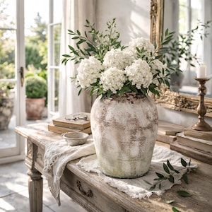 Puede incluir: Un jarrón grande de cerámica envejecida lleno de flores de hortensia blancas y follaje verde se encuentra sobre una mesa de madera desgastada. Un cuenco pequeño, libros y un candelabro también están sobre la mesa, creando una exhibición rústica y elegante.
