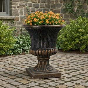 Könnte beinhalten: Ein großer, verzierter Außenblumentopf in Schwarz und Bronze, gefüllt mit orangefarbenen und gelben Blumen. Der Topf hat einen geriffelten Körper und steht auf einem quadratischen Sockel, ideal für Garten oder Terrasse.