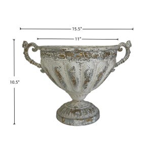 May include: A distressed white and gold-toned decorative urn, 15.5 inches wide, 11 inches in diameter, and 10.5 inches tall. The urn features ornate handles and a pedestal base, suitable for home decor or floral arrangements.