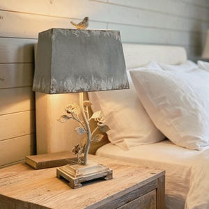 May include: A decorative bedside lamp with a gray rectangular lampshade and a bird figurine on top. The lamp base features a floral design. The lamp is placed on a wooden nightstand next to a bed with white linens.