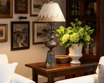Puede incluir: Una lámpara de mesa de estilo vintage con una base azul y dorada desgastada y una pantalla crema. La lámpara está sobre una mesa de madera oscura, junto a un jarrón blanco lleno de flores verdes. La habitación presenta obras de arte enmarcadas.