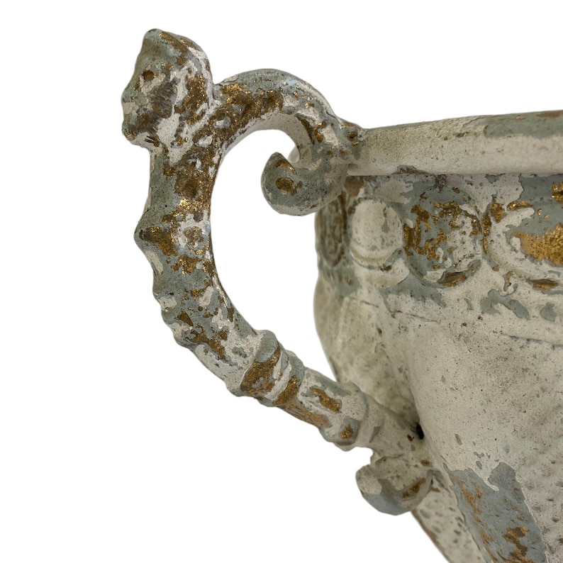 May include: Close-up of a decorative, antique-style urn with a handle. The urn is painted in a distressed style with a white base, light blue accents, and gold highlights. The handle has a detailed, ornate design.