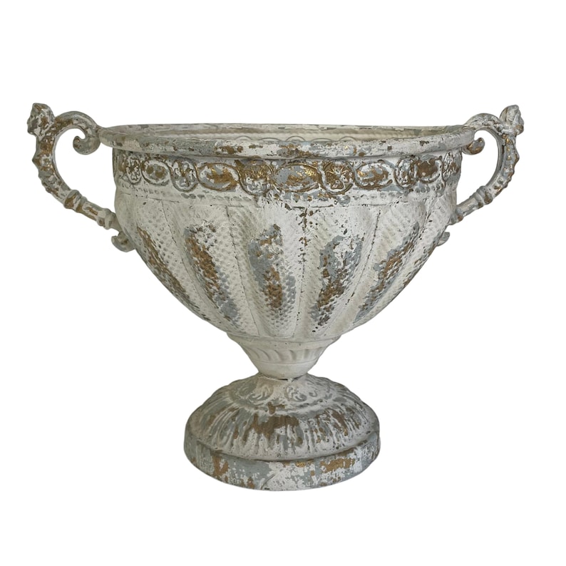 May include: A distressed white and gold decorative urn with two ornate handles. The urn has a textured, ribbed body and sits on a pedestal base. The aged finish gives it a vintage, antique appearance, suitable for home decor.