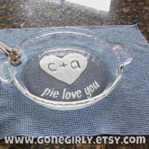 May include: Clear glass pie dish with etched text and a heart design. The heart contains the letters "C+A". The words "pie love you" are etched below the heart. The Etsy shop URL is visible at the bottom.