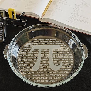 May include: A clear glass pie dish with the mathematical symbol pi and a series of numbers etched on the bottom. A calculator, glasses, and an open book are next to the dish. The dish is on a black cloth.