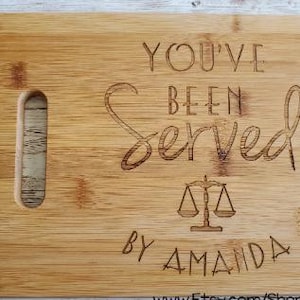 May include: A rectangular bamboo cutting board with a handle cutout. The board is engraved with the words "YOU'VE BEEN SERVED" above a scale of justice symbol and the name "AMANDA".