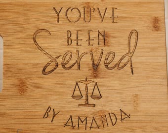 May include: A rectangular light brown wooden cutting board featuring a laser-engraved design. The text reads YOU'VE BEEN SERVED, followed by an icon of a scale of justice, and the name AMANDA at the bottom. The wood grain is clearly visible.