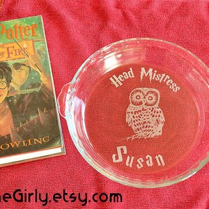 May include: A clear glass pie dish with the words "Head Mistress Susan" and an owl etched on the red surface. Beside it is a copy of "Harry Potter and the Goblet of Fire" by J.K. Rowling.