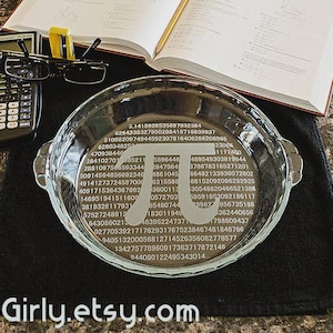 May include: A clear glass pie dish with the mathematical symbol pi and the digits of pi etched into the bottom. A calculator, glasses, and an open book are nearby. The dish sits on a black cloth.
