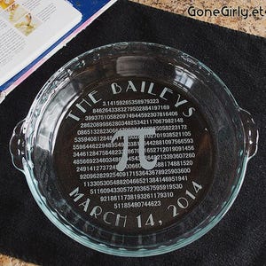 May include: Clear glass pie dish with etched text. The text includes "The Baileys", the number pi, and "March 14, 2014". The dish has handles and sits on a black surface.