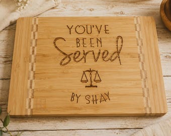 May include: A rectangular bamboo cutting board with a light wood grain and vertical checkered side panels. The center is laser-engraved with the text "YOU'VE BEEN SERVED" above a graphic of a scale of justice and the text "BY SHAY".