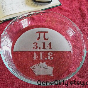 May include: Clear glass pie dish with etched design. The design includes the Pi symbol, the numbers 3.14 and 41.8, and a pie slice graphic. The dish has scalloped edges and handles. The background is a red fabric.