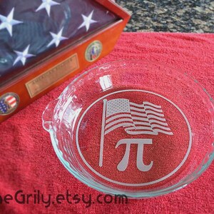 May include: Clear glass pie dish with an etched design featuring the American flag and the pi symbol. The dish is on a red towel, with a wooden flag display case in the background.