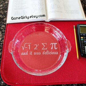 May include: A clear glass pie dish with etched mathematical symbols and the phrase "and it was delicious." The dish sits on a red mat, with a textbook, pencil, and calculator in the background. The Etsy shop name is visible.