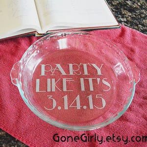 May include: A clear glass pie dish with the text "PARTY LIKE IT'S 3.14.15" etched in the center. The pie dish sits on a red cloth, with a cookbook open in the background. The Etsy shop name is visible.