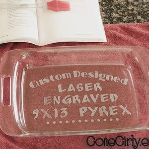 May include: A clear glass 9x13 inch Pyrex baking dish with laser-engraved text that reads "Custom Designed Laser Engraved 9x13 Pyrex." The dish sits on a red towel, with an open cookbook and a red silicone brush in the background.