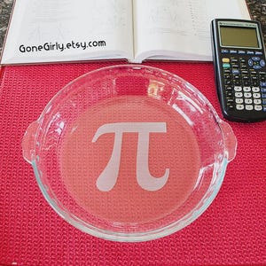 May include: A clear glass pie dish with a large white "pi" symbol etched in the center, set on a red textured mat. A calculator and an open book are in the background. The Etsy shop name "GoneGirly.etsy.com" is visible.