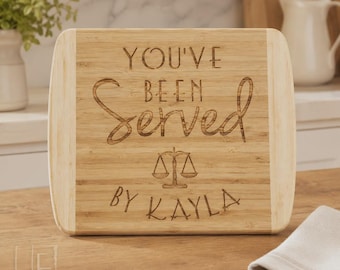 May include: A rectangular bamboo cutting board with rounded corners featuring the laser-engraved text "YOU'VE BEEN Served" above a graphic of a scale of justice and the name "BY KAYLA" at the bottom, showcasing a light wood grain finish.