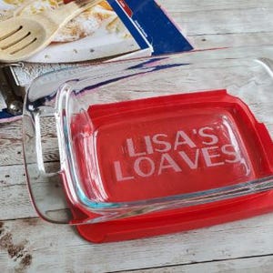 May include: A clear glass baking dish with a red lid. The lid has the text "LISA'S LOAVES" in white. A wooden spoon and a cookbook are in the background. The dish is on a light-colored wooden surface.