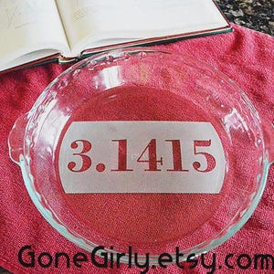 May include: A clear glass pie dish with a white stencil of the mathematical constant "3.1415" on a red cloth. The pie dish has a scalloped edge and two handles. An open book is in the background.