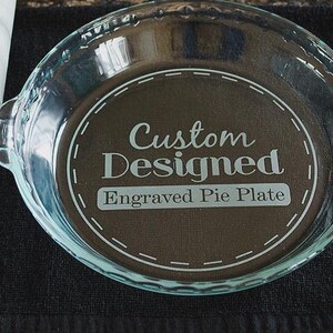 May include: Clear glass pie plate with the words "Custom Designed Engraved Pie Plate" in white. A marble rolling pin is on the left. The pie plate is on a black surface.