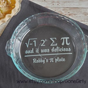 May include: Clear glass pie plate with a fluted edge and handles. The plate features the text "√-1 2³ Σ π and it was delicious Robby's π plate" in white. A partially visible pie sits in the background.