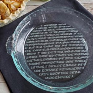 May include: Clear glass pie dish with a decorative rim and handles. The bottom of the dish is etched with the digits of Pi. A partially visible apple pie sits nearby on a dark gray cloth.