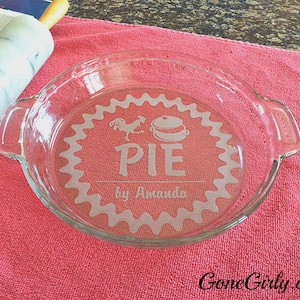 May include: A clear glass pie dish with etched design, including a rooster and pie graphic, and the text "PIE by Amanda." The dish sits on a pink towel, with a rolling pin and cookbook visible.