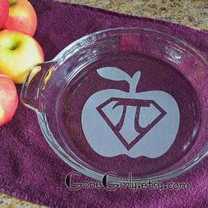 May include: A clear glass pie dish with a decorative apple design featuring the pi symbol inside. The dish is surrounded by red and yellow apples on a purple surface.