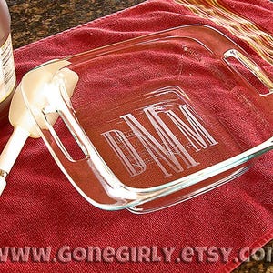 May include: A clear glass baking dish with engraved initials, resting on a red towel. A white spatula and a bottle of honey are also in the image. The dish has handles and is rectangular in shape.