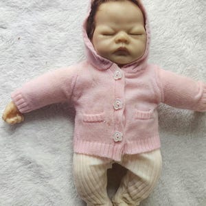 Può includere: Una bambola realistica con un maglione con cappuccio rosa chiaro con tre bottoni bianchi a forma di fiore e tasche. La bambola indossa anche pantaloni a coste color crema e ha i capelli castani.