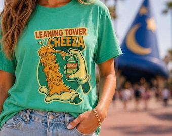 May include: A teal t-shirt with the text "LEANING TOWER of CHEEZA" and a cartoon graphic of a cheese tower being sprayed with "Cheddar Whizzy" cheese. The shirt is worn with blue jeans.