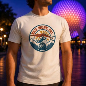 May include: Cream-colored t-shirt featuring a circular graphic with the text "NICE WORK, PAL." The graphic depicts a hang glider, clouds, and a geometric sphere. The design is in shades of orange, blue, and yellow.