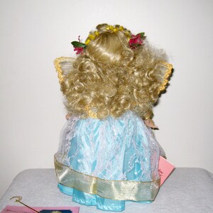 May include: A porcelain doll with curly blonde hair, wearing a light blue dress with white lace overlay and gold trim. The doll has a floral crown and small wings. A pink tag is visible.