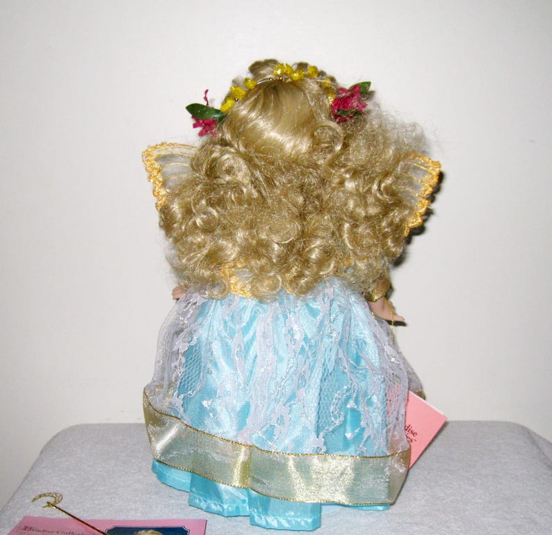 May include: A porcelain doll with curly blonde hair, wearing a light blue dress with white lace overlay and gold trim. The doll has a floral crown and small wings. A pink tag is visible.