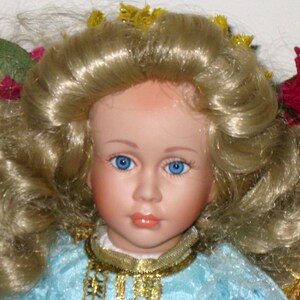 May include: A porcelain doll with blonde, curly hair and bright blue eyes. The doll is wearing a light blue dress with a gold necklace. Decorative flowers and leaves adorn the doll's hair. The doll has a serene expression.