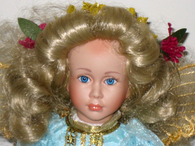 May include: A porcelain doll with blonde, curly hair and bright blue eyes. The doll is wearing a light blue dress with a gold necklace. Decorative flowers and leaves adorn the doll's hair. The doll has a serene expression.
