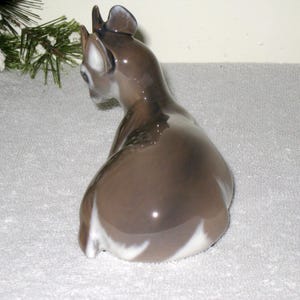May include: A porcelain figurine of a fawn sitting on its haunches. The fawn is brown with white markings on its belly and legs. The figurine is glazed and has a glossy finish.