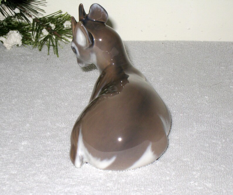 May include: A porcelain figurine of a fawn sitting on its haunches. The fawn is brown with white markings on its belly and legs. The figurine is glazed and has a glossy finish.