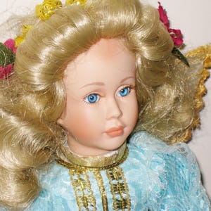 May include: A porcelain doll with blonde, curly hair and blue eyes. The doll wears a light blue dress with gold trim and a gold necklace. Floral accents are in the doll's hair.